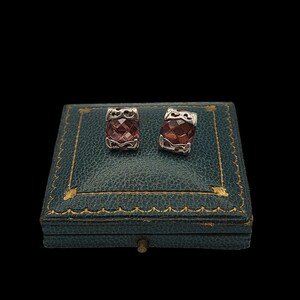 May include: Two silver earrings with faceted, reddish-brown gemstones. They are presented on a vintage teal jewellery box with gold trim. The box is closed and set against a black background.