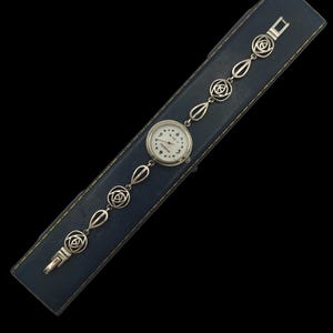 May include: A vintage silver-toned wristwatch with a white face and black numerals, resting on a blue leather case. The watch band features a series of decorative links with floral and geometric designs. The watch is a classic accessory.
