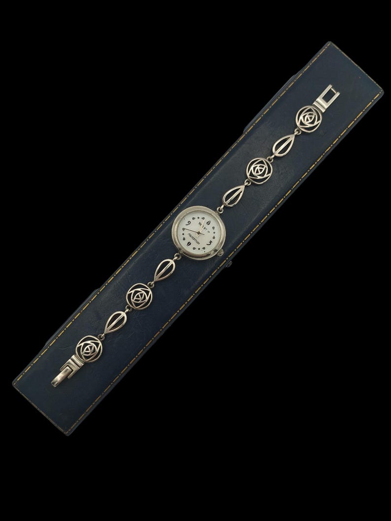 May include: A vintage silver-toned wristwatch with a white face and black numerals, resting on a blue leather case. The watch band features a series of decorative links with floral and geometric designs. The watch is a classic accessory.