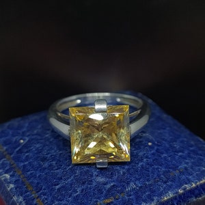 Op de afbeelding: Een zilveren ring met een grote, vierkante, gele edelsteen in het midden.
