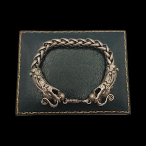 May include: A silver bracelet with two dragon heads on the clasp. The bracelet is displayed on a green velvet background.