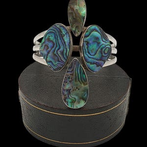 Puede incluir: Una pulsera de plata con cuatro adornos de concha de abulón en tonos azules, verdes y morados. La pulsera tiene un diseño único con tres bandas paralelas. Las conchas de abulón tienen forma de lágrima y ovalada.