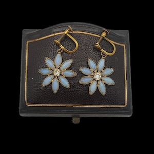 May include: A pair of vintage screw-back earrings. Each earring features a flower design with light blue petals and a central clear gemstone. The earrings have a gold-tone metal setting and are displayed on a black jewellery box.
