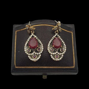 May include: A pair of silver earrings with a teardrop-shaped red gemstone surrounded by smaller clear gemstones. The earrings are in a small box.