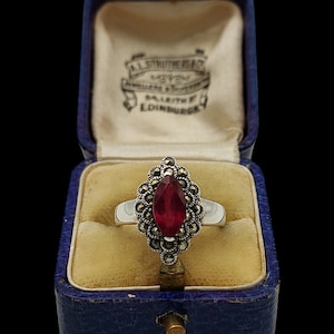 May include: A silver ring with a large, oval, deep red gemstone, surrounded by smaller, round, silver-colored stones. The ring is displayed in a vintage blue jewelry box with a yellow interior. The box has text that reads "A.L. STRUTHER & CO. LEITH & EDINBURGH."