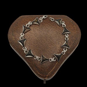 May include: Silver bracelet featuring triangular black details. The bracelet is made up of multiple triangular shapes joined by small silver links. The bracelet is presented on a brown, heart-shaped box against a black backdrop.