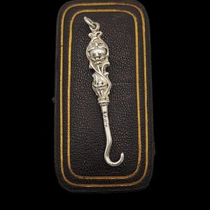 Puede incluir: Un amuleto de plata en forma de gancho con un diseño floral. El amuleto está en una caja de terciopelo negro con ribetes dorados.