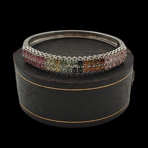Könnte beinhalten: Ein silbernes Armband mit zwei Reihen farbiger Edelsteine, darunter rosa, grüne, gelbe und braune Steine. Das Armband liegt auf einer schwarzen, runden Schatulle mit goldenem Rand. Der Hintergrund ist schwarz.