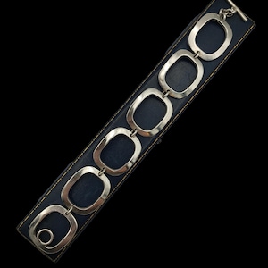 May include: A silver bracelet featuring six linked, open rectangular shapes. The bracelet is presented on a dark blue jewellery box against a black backdrop. It has a T-bar clasp. The bracelet is approximately 19 cm long.