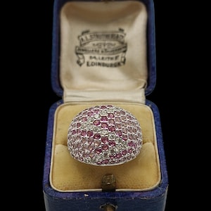 May include: A silver ring with a domed surface, encrusted with small pink and clear gemstones. The ring is displayed in a vintage blue jewelry box with a cream-colored interior and a gold clasp. The box has text on the inside of the lid.