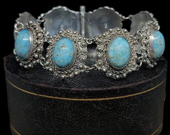 Op de afbeelding: Een zilveren armband met vijf turquoise stenen gezet in sierlijke zilveren frames. De armband ligt op een zwarte fluwelen doos.