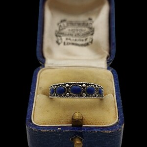 May include: A silver band ring featuring five small, oval-shaped, deep blue stones set in a row. The metal band has a textured, beaded border design along the top and bottom edges. The ring is displayed inside a blue, hinged jewellery box.