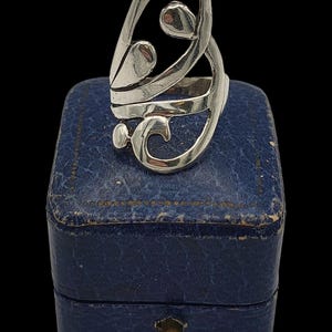 Könnte beinhalten: Ein silberfarbener Ring mit einem abstrakten Design, das geschwungene und ovale Formen aufweist. Der Ring wird auf einer blauen Vintage-Schmuckschatulle vor schwarzem Hintergrund präsentiert. Das Design des Rings ist offen und luftig.