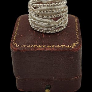 May include: A silver ring with a textured design, featuring multiple bands with a rope-like pattern. The ring is displayed on a small, antique, brown jewelry box with gold trim. The background is black.