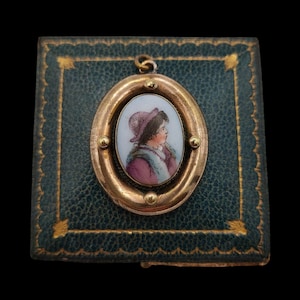 May include: An oval gold pendant featuring a hand-painted enamel portrait of a person wearing a purple hat and coat with a white fur collar. The pendant is set against a square, dark green textured box with gold decorative border detailing.