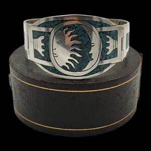 May include: A silver cuff bracelet with turquoise inlay and a central oval design featuring a sun and moon motif. The bracelet is displayed on a dark, textured surface, showcasing its intricate details and craftsmanship.