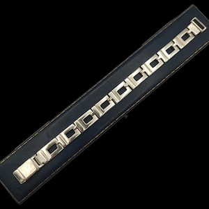 May include: A silver-toned bracelet with a geometric design, presented in a dark blue rectangular box. The bracelet features a series of connected rectangular links. The box has a textured appearance and a gold-colored trim.