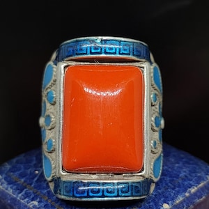 Puede incluir: Un anillo de plata con una piedra roja engastada en un marco de esmalte azul. El marco está decorado con un patrón geométrico.