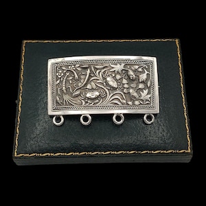 Puede incluir: Broche rectangular de plata con intrincados diseños florales y acuáticos. El broche tiene tres pequeños lazos en la parte inferior. Se muestra sobre una caja verde oscuro con ribete dorado.