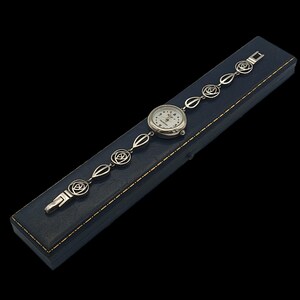 May include: A silver-toned wristwatch with a white face and black numerals rests on a dark blue box. The watch band features a series of rose-shaped links. The watch is a classic accessory.