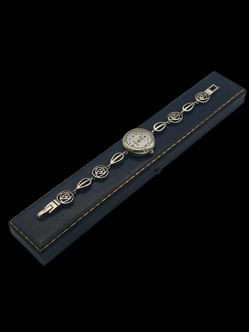 May include: A silver-toned wristwatch with a white face and black numerals rests on a dark blue box. The watch band features a series of rose-shaped links. The watch is a classic accessory.