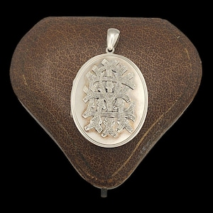 May include: Silver locket with an intricate design on the front. The locket is in a brown leather case.