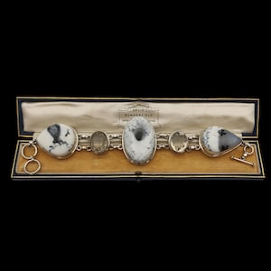 Puede incluir: Un brazalete de plata con tres piedras grandes ovaladas de color blanco y gris y dos piedras más pequeñas transparentes. El brazalete está en una caja forrada de terciopelo con el nombre "David Summerfield, Nueva York, Londres" impreso en el interior.