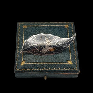 May include: A silver-toned leaf-shaped brooch with a detailed design of a figure. The brooch is set on a vintage teal jewelry box with gold trim. The brooch has a detailed design of a figure.