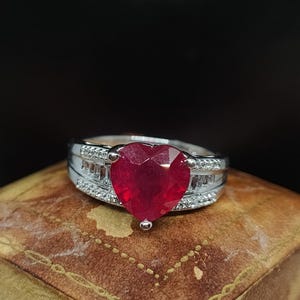 Może przedstawiać: Srebrny pierścionek z rubinem w kształcie serca otoczonym mniejszymi diamentami.