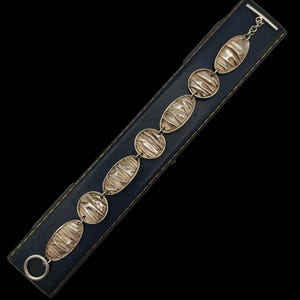 May include: Silver-toned bracelet with oval and circular textured links. The bracelet is displayed on a dark blue rectangular box with a black background. The bracelet has a toggle clasp closure.