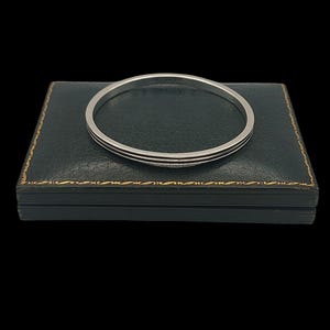 Op de afbeelding: Een zilveren armband met twee dunne, parallelle groeven. De armband ligt op een donkergroene doos met gouden rand. De achtergrond is zwart, waardoor het sieraad wordt benadrukt.
