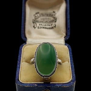 May include: A silver ring featuring a large, smooth, oval-shaped green stone set in a decorative metal bezel. The ring is displayed inside a vintage blue velvet-lined jewelry box with the text A. STRUTHERS JEWELLER & SILVERSMITH 34 LEITH EDINBURGH.