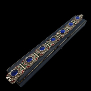 Puede incluir: Un brazalete de plata con detalles en esmalte azul, rojo y verde y piedras de vidrio azul. El brazalete está en una caja de terciopelo azul.