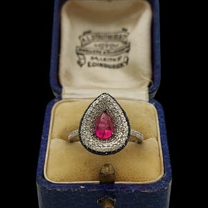 Könnte beinhalten: Silberring mit einem tropfenförmigen, rubinroten Edelstein. Der Stein ist von einem Kranz aus kleinen, klaren Steinen und einem schwarzen Rand umgeben. Der Ring wird in einer blauen Schmuckschatulle präsentiert.