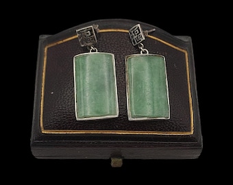 Può includere: Un paio di orecchini rettangolari in giada verde con montature in metallo color argento e top decorativi. Gli orecchini sono esposti in un portagioie nero con finiture dorate.