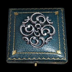 May include: A silver filigree brooch in a green velvet box. The brooch is a circular design with intricate swirls and curves.
