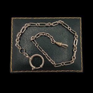 May include: A silver-toned metal watch chain featuring a mix of textured and smooth elongated links. The chain includes a large circular spring ring clasp at one end and a tapered metal fob attachment at the other, creating a classic jewellery piece.