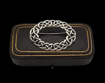 Pode incluir: Um broche de nó celta de prata em forma de círculo, apresentado em uma caixa de veludo preto com guarnições douradas.