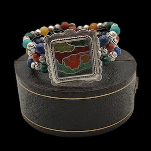 May include: A multi-strand bracelet with colourful beads in shades of blue, green, red, yellow, and turquoise. The bracelet features a square, silver-toned centrepiece with a mosaic design in shades of red, green, and brown. The bracelet is resting on a black box.