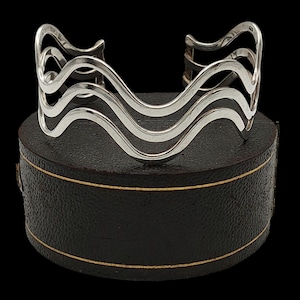 May include: A silver cuff bracelet with a wave design. The bracelet features three wavy bands that flow across the wrist. The jewelry is displayed on a black box with a gold trim.