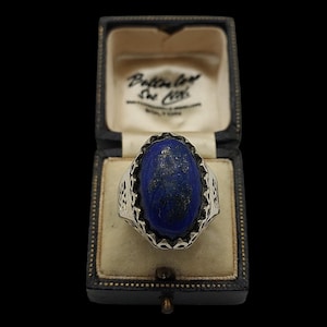 May include: A silver ring featuring a large, oval-shaped, deep blue lapis lazuli stone with gold-colored flecks. The metal band has an intricate, cut-out filigree pattern along the sides. The ring is displayed inside a dark, hinged jewelry box.