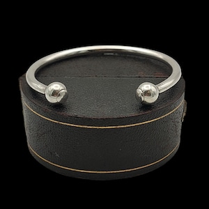 May include: A silver cuff bracelet with a smooth, rounded design. The bracelet has a simple, open design with a ball at each end. The bracelet is resting on a black, leather-effect box with gold trim.