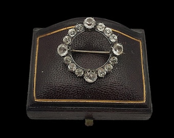 Könnte beinhalten: Eine silberfarbene Vintage-Brosche mit kreisförmigem Design, besetzt mit zahlreichen klaren, funkelnden Edelsteinen. Die Brosche wird auf einer dunkelbraunen Schmuckschatulle mit goldenem Rand vor schwarzem Hintergrund präsentiert.