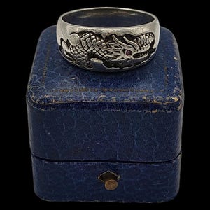 Puede incluir: Anillo plateado con un diseño detallado de dragón. El dragón tiene un ojo de gema roja. El anillo se exhibe en una pequeña caja de joyería azul vintage con un cierre de latón. El fondo es negro.