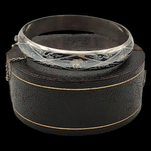 May include: A silver hinged bangle with detailed black enamel patterns. The bracelet is circular and rests on a dark brown, round jewellery box with gold edging. The design features a repeating pattern.