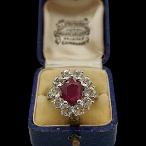 May include: A silver ring featuring a central oval ruby, encircled by a cluster of smaller, clear gemstones. The ring is presented in a vintage blue jewellery box with a cream interior.
