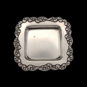 May include: A square, silver-coloured tray with an ornate border. The tray has a slightly raised edge and a smooth, reflective surface. The border features a repeating scroll design. The tray is set against a black background.