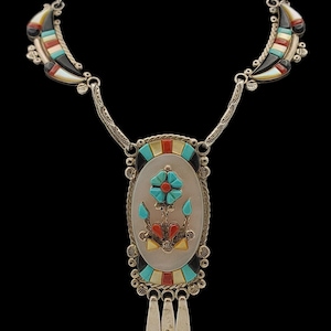 May include: Silver necklace with a central oval pendant featuring a turquoise flower design. The pendant is surrounded by colorful inlaid stones. Crescent-shaped accents with inlaid stones are on each side of the necklace. The necklace has three dangling silver pendants.