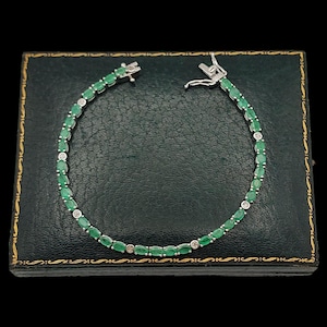 Puede incluir: Una pulsera de plata con esmeraldas ovaladas y pequeños diamantes. La pulsera se muestra sobre una superficie verde oscuro texturizada, con un cierre en la parte superior. Las piedras preciosas están dispuestas en línea, creando un diseño clásico y elegante.