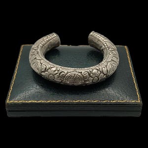 Puede incluir: Un brazalete plateado con intrincados diseños florales y geométricos. El brazalete abierto se exhibe sobre una caja verde oscuro con ribete dorado, sobre un fondo negro. El diseño del brazalete incluye tallas detalladas.